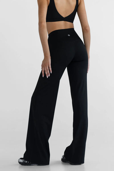A lifestyle studio portrait capturing a model posing from the side in the black Tall Sculpt Wide Leg Pants and matching sports bra. The image highlights the flexibility, 100% squat-proof coverage, and ultra-soft texture of the nylon-spandex blend fabric.