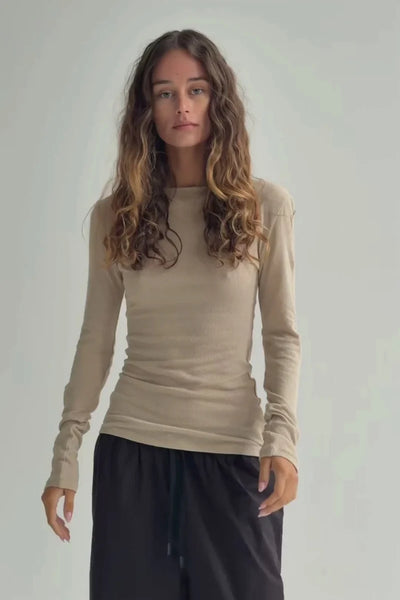 Front view of a model with long wavy hair wearing the lightweight, semi-sheer Zulu & Zephyr Rib Hemp Jersey Long Sleeve Top in husk beige styled with black trousers.