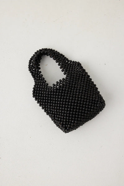 A direct overhead studio flat lay of the black beaded bucket bag resting on a clean white surface, showcasing its minimalist, unlined structure and tightly woven geometric grid of matte wooden beads.