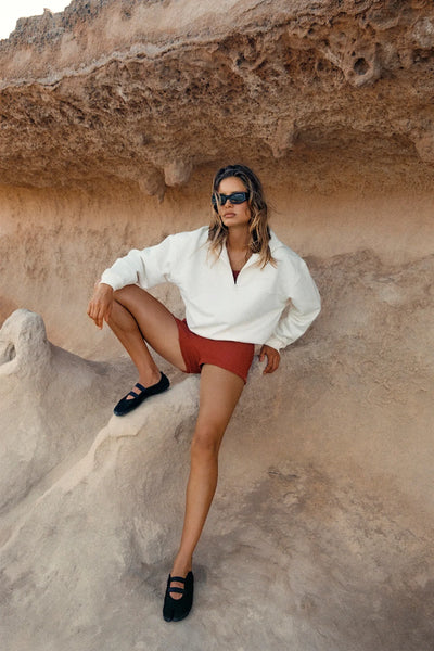 Campaign photo of a model crouching on rocks wearing the cream quarter-zip Zulu & Zephyr Sport Sweat Shirt Fleece, rust shorts, and sunglasses.