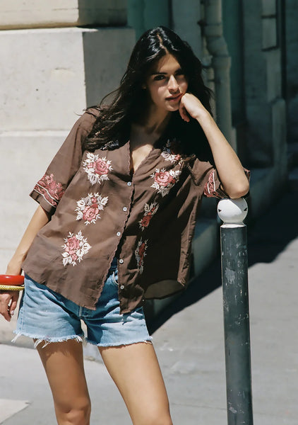 Outdoor campaign shot of a model with dark hair leaning against a street post while wearing the relaxed-fit brown Auguste Rosalie Blouse. The blouse is casually styled open over high-waisted denim shorts with a frayed hem, accessorized with bold red and gold stacked bangles.