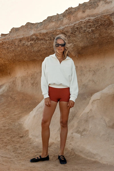 Outdoor campaign shot of a model wearing the Zulu & Zephyr Sport Sweat Shirt Fleece in Cream, styled with rust-red bike shorts and black sunglasses against rocky terrain.