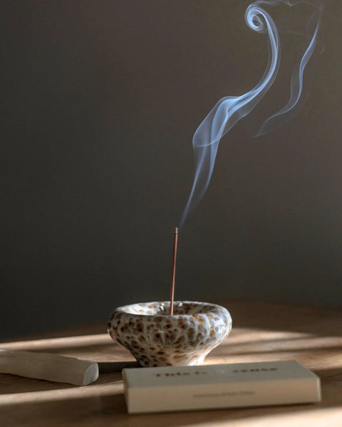 Mood shot of a lit incense stick burning in the Gentle Habits Vessel Incense Holder in Seafoam with smoke rising against a dark, warm-lit background.