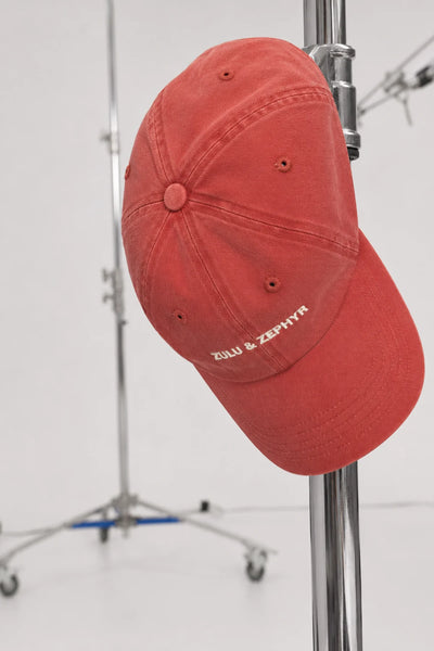 Close-up studio view of the red Zulu & Zephyr Canvas Cap hanging on a metal stand, highlighting the curved brim and white logo embroidery.
