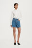 Full-length studio front view of a model standing with one hand in the pocket, wearing the high-rise blue denim shorts, white button-down shirt, and black heels.