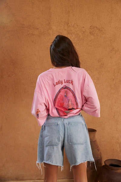 Back view of a model wearing Sabbi The Very Oversized Lucky Mermaid Tee in Bright Pink styled with light-wash denim shorts, showing the "Lady Luck" artwork.