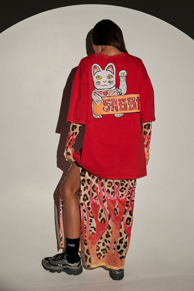 Full-length rear view of a model in studio lighting wearing the red SABBI Very Oversized Champagne Tattoo Tee with the "SABBI" lucky cat back art, styled with a printed maxi skirt and black sneakers.