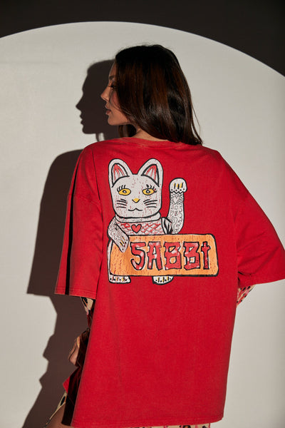Rear half-body studio portrait of a model looking over her shoulder wearing the red oversized SABBI graphic t-shirt with the illustrated lucky cat back print.