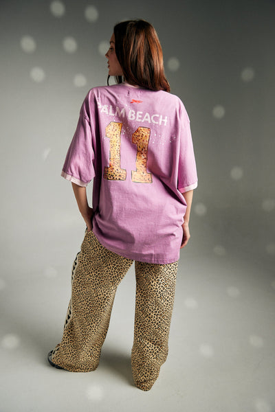 A full-length lifestyle campaign photograph from the back, showing a model walking away to highlight the large, statement varsity lettering and running cheetah artwork printed across the upper back panel of the pink tee.