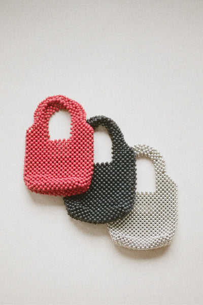 A soft-focus artistic group layout showcasing three iterations of the petite purse one coral red, one black, and one pearlescent silver arranged together to illustrate the distinct profile of the tightly beaded silhouette.