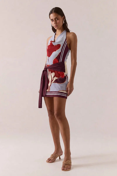 Three-quarter full-length front view of a model posing in the Vere Kalea Blue Bloom mini dress featuring an asymmetric cowl neck, waist tie detail, and floral stretch mesh pattern.
