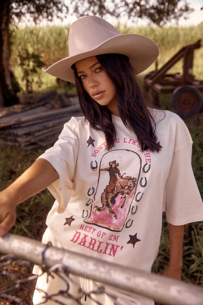 An outdoor country lifestyle campaign image capturing a model from the side in a rustic outdoor setting, wearing the slouchy marshmallow white Darlin Tee. The portrait highlights the relaxed sleeve drape, the worn-in charm of the enzyme wash, and the distinct western-inspired graphics printed down the sleeve.