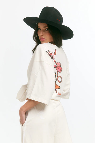 A full-length studio product shot from the back on a model styling the marshmallow white oversized tee as a t-shirt dress. The image showcases the distressed enzyme-washed texture of the 100% cotton fabric and the large hand-designed western graphic layout spanning across the back panel.