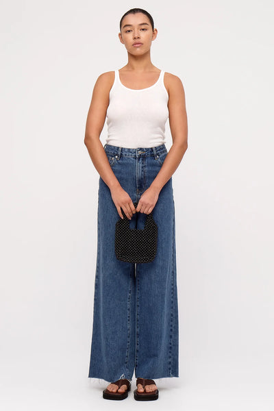 A full-length studio shot of a model wearing wide-leg denim and a white tank top, holding the black wooden beaded bucket bag in front of her to provide a clear sense of its compact, evening-ready scale.