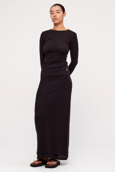 Three-quarter view of a model with slicked-back hair wearing the slim-fit Zulu & Zephyr Rib Hemp Jersey Long Sleeve Top in black, styled with a matching black maxi skirt.