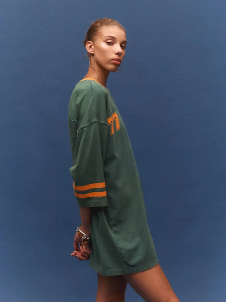 A three-quarter studio profile view of a model facing right against a solid blue background. She is wearing the stone-washed green tee dress, highlighting the relaxed fit, dropped shoulder, and the vintage-style orange stripes on the three-quarter sleeve.
