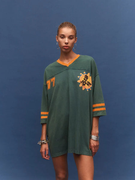 A front-on studio portrait of the model standing straight against a solid blue backdrop. The image showcases the V-neckline with orange trim, the oversized jersey construction, the "77" graphic on the right shoulder, and the vibrant Studio Zube sun graphic on the left chest.