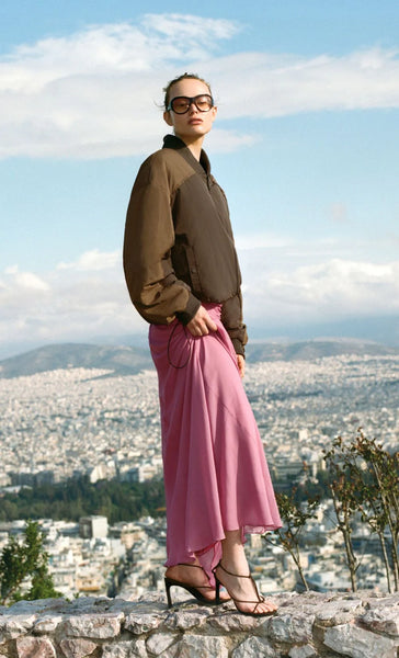 Outdoor editorial campaign image of a model wearing the Bec + Bridge Laney Jacket in Choc/Coffee styled with a flowy pink midi skirt and sunglasses against a scenic city overlook.