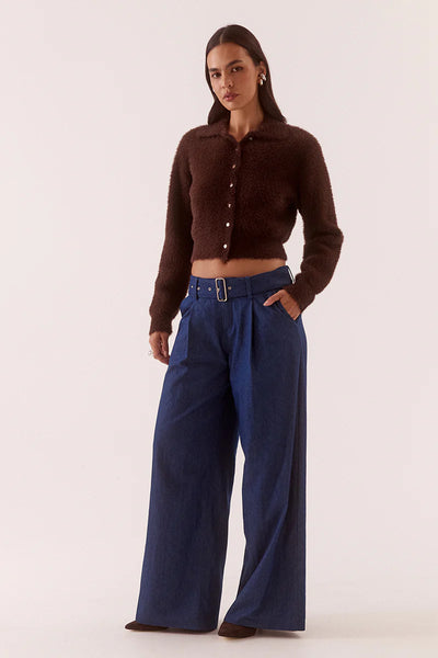 A full-length studio shot of the model posing at a slight three-quarter angle. The framing captures the rum raisin brown long-sleeve knitwear piece styled buttoned up over a bare midriff, highlighting the texture of the feather-light yarn, the ribbed sleeve cuffs, and the pairing with belted dark blue jeans.
