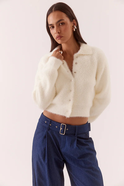 A full-length studio shot of the model standing at a three-quarter angle looking forward. The image isolates the left side of the chalk-colored 100% nylon cardigan, highlighting the natural drape of the plush, long sleeves, the short buttoned front placket completely closed, and the relaxed pairing of the feather-light texture with pleated denim trousers.