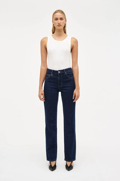 Full-length studio portrait of a model dressed in a coordinated dark indigo denim set, featuring the Neuw Freja Straight jeans and the matching buttoned Stella Jacket over a white top. The clean, modern double-denim look is styled with black heels and gold hoop earrings against a white background.
