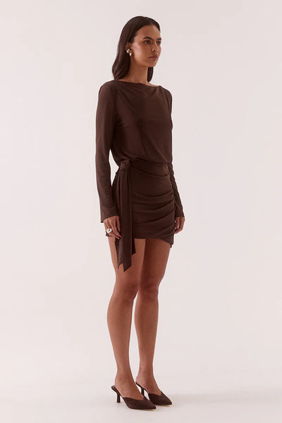 A full-length studio shot of the model posing from a right-side profile angle. The framing captures the sleek line of the long sleeves and the architectural fluid tuck details at the hip, with the fabric tie hanging elegantly from the waist.