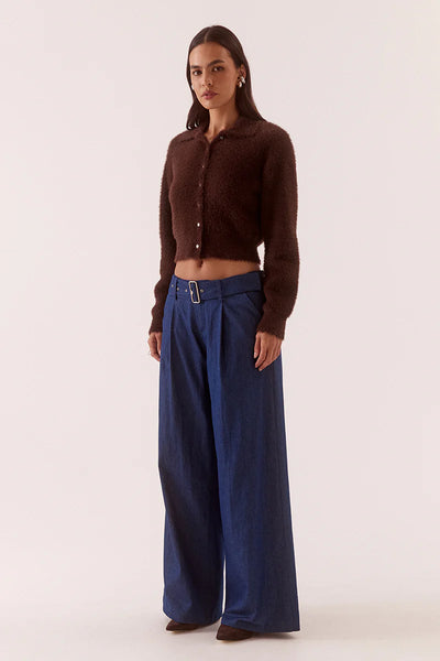 A full-length front-on studio portrait of the model posing with one hand resting inside her trouser pocket. The image provides a clear view of the cardigan's front architecture, showcasing the sharp pointed collar, the short placket with small light-contrast buttons, and the close, body-skimming fit of the 100% nylon knit fabric.