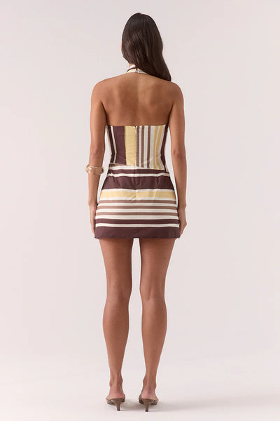 A full-length studio shot from behind a model wearing the Sovere Sonali Skortsuit in the signature Banoffee brown, yellow, and white striped print. The image highlights the open-back halter neck silhouette, the fitted back bodice with a centered invisible zipper, and the clean mini length cut of the skort's back hemline.