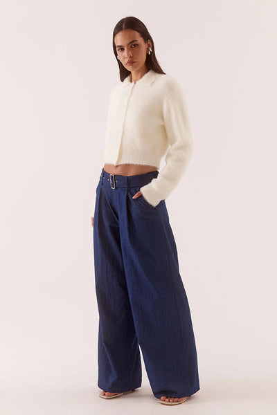 A close-up mid-shot studio portrait focusing on the upper torso and delicate textile detail of the chalk white top. The cardigan is styled slightly open at the top, revealing silver metallic buttons and an unbuttoned knit collar, while highlighting the ultra-soft, eyelash-style fluffy yarn texture against the model's hand and statement silver ear cuffs.