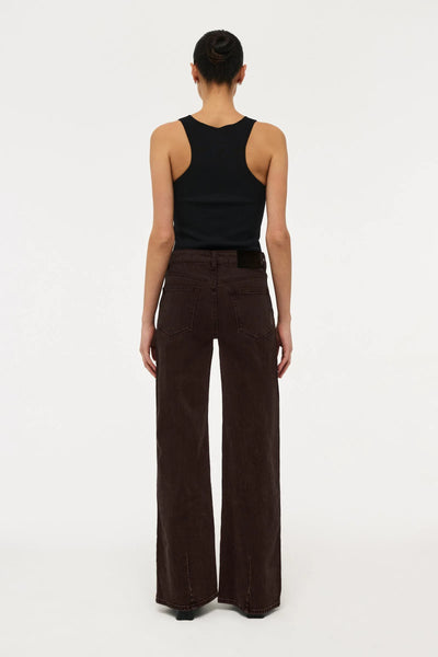 Rear full-length view of a model wearing Neuw Eva Wide jeans in Sable brown, featuring back patch pockets, black leather brand patch, and wide-leg hem paired with a black ribbed tank top.