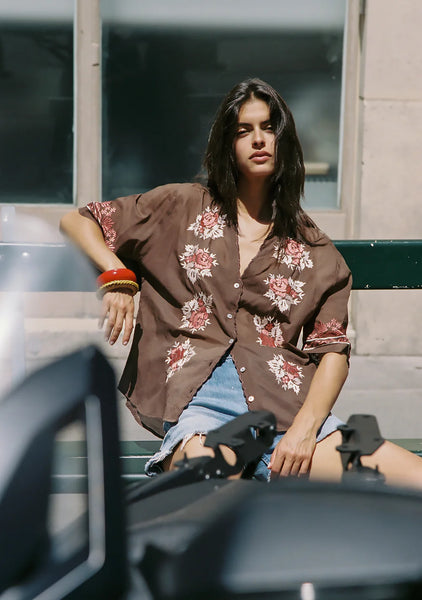 A sun-drenched outdoor campaign lifestyle shot of a model sitting on a green bench, wearing the lightweight cotton voile Auguste Rosalie Blouse in Cocoa. The image captures the semi-sheer finish of the brown fabric and highlights the intricate hand-embroidered bohemian details paired with casual denim shorts.