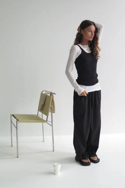 Full-length lifestyle shot of a model wearing the white Zulu & Zephyr Rib Hemp Jersey Long Sleeve Top layered under a black cami top with wide-leg trousers.