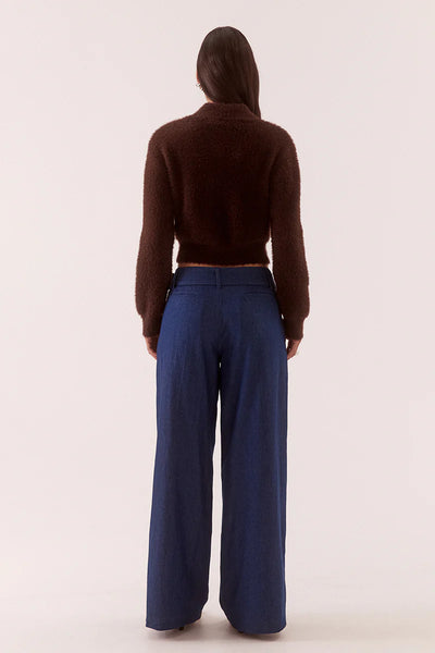 A full-length rear view photograph of a model standing straight, facing away from the camera against an off-white studio background. She is wearing the plush rum raisin dark brown fluffy knit cardigan, illustrating the fully fashioned construction across the back, subtle dropped shoulder seams, and a straight, cropped fitted hemline paired with high-waisted wide-leg dark denim trousers.