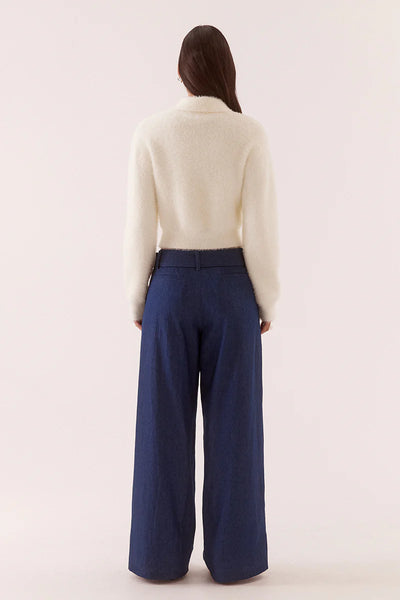 A full-length front-on studio portrait of the model standing straight against an isolated pale backdrop. The shot frames the complete outfit layout, showcasing the chalk white Fenna knit buttoned mid-way to reveal a hint of midriff, emphasizing the symmetrical rib hem, matching rib sleeve cuffs, sharp pointed collar, and the contrast with tailored, belted wide-leg blue jeans.