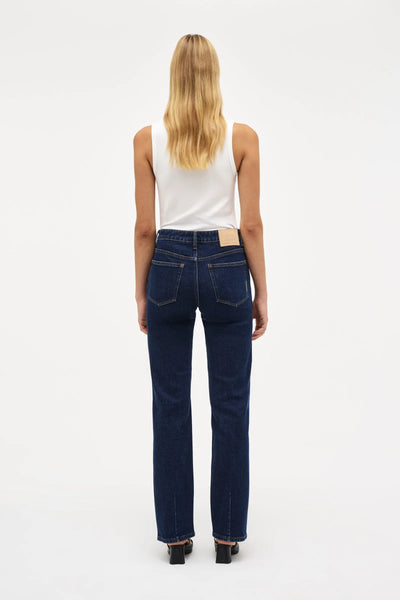 Full-length studio back-view of a blonde model standing against a white background, wearing the Neuw Freja Straight jeans in dark indigo paired with a white ribbed tank top and black low heels. The shot highlights the flattering fit of the back pockets and the straight, stovepipe fall of the legs.