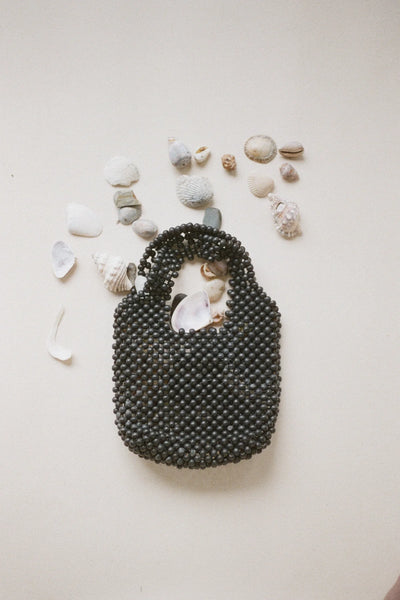 A high-angle shot featuring two petite beaded bags cast in soft diagonal light, displaying the black version alongside a warm brown counterpart to highlight the subtle texture differences and integrated clutch handles.