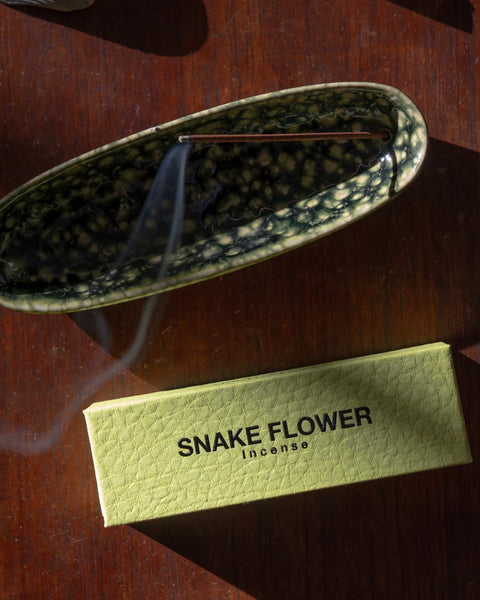 Overhead view of the Gentle Habits Collector Ceramic Holder in mottled green glazed ceramic with a lit incense stick resting inside next to a box of Snake Flower Incense.