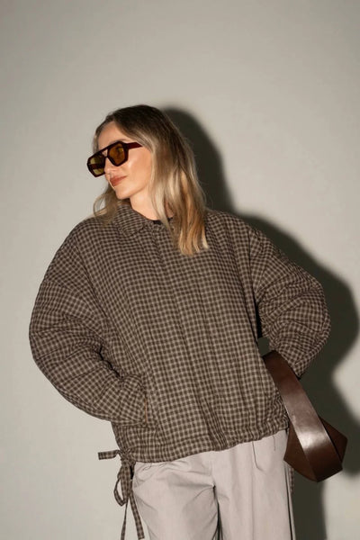 A stylized shot of the jacket paired with light grey trousers and a large brown leather bag. The model wears yellow-tinted sunglasses, emphasizing a modern, urban aesthetic.