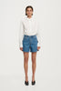Full-length studio front view of a model wearing the high-rise blue denim shorts paired with a tucked-in crisp white button-down shirt and black heeled mules.