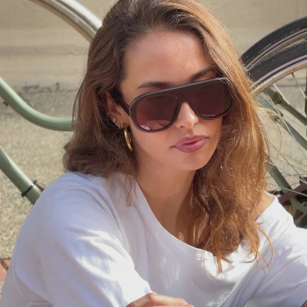 Casual outdoor portrait of a brunette model sitting in a white top, wearing oversized translucent milky coco aviator sunglasses.