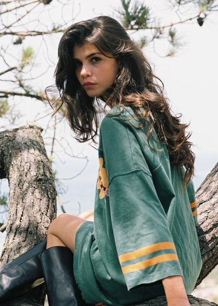 A casual, over-the-shoulder outdoor photograph of the model perched in a pine tree. Looking back toward the camera, the shot highlights the roomy back construction of the relaxed jersey dress, the texture of the faded stonewash fabric, and the orange stripes on the sleeve.