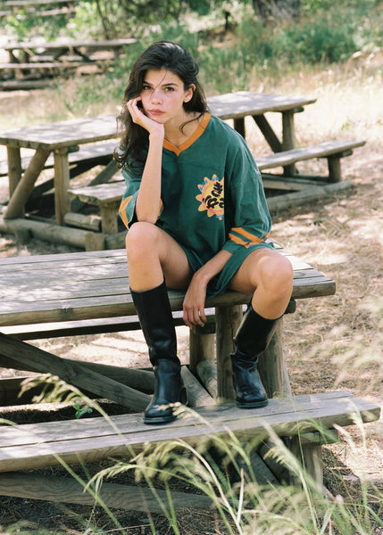 An outdoor lifestyle shot of a model sitting on a rustic wooden picnic table in a sunny, wooded environment. She wears the relaxed 100% cotton green tea dress paired with knee-high black leather boots, leaning forward with her chin resting in her hand.