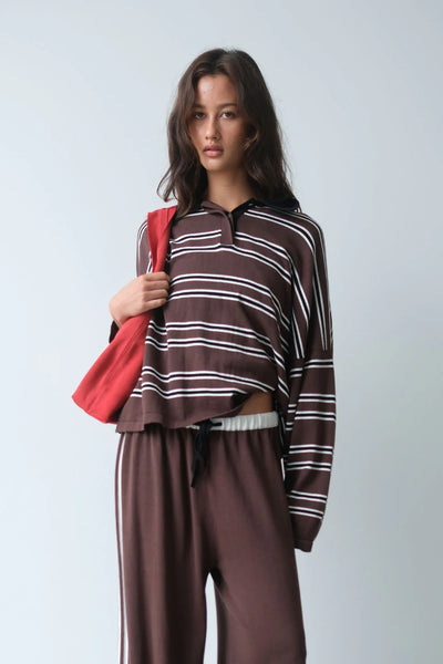 A campaign-style studio portrait of a model wearing the oversized espresso stripe Rayne polo knit jersey. She has a red canvas shoulder bag slung over her arm and is paired with matching espresso brown track pants featuring white side stripes, creating a sporty, casual layering look.