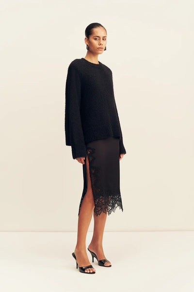 Three-quarter standing pose of a model styled in the black Shona Joy Korine shoulder-padded boucle knit jumper with a side-split lace trim midi skirt.