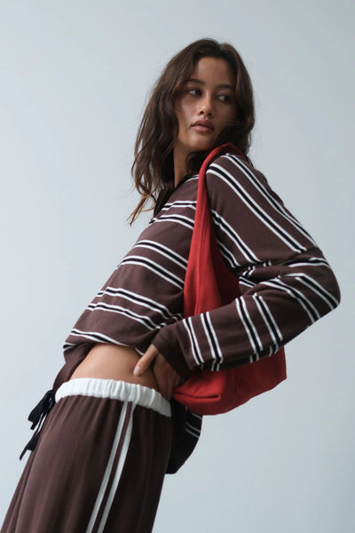 An expressive, low-angle lifestyle shot featuring the model leaning back dynamically against a clean gray background. The shot captures the relaxed drape of the striped long sleeves and showcases a glimpse of the contrasting white waistband on the coordinating lounge pants.