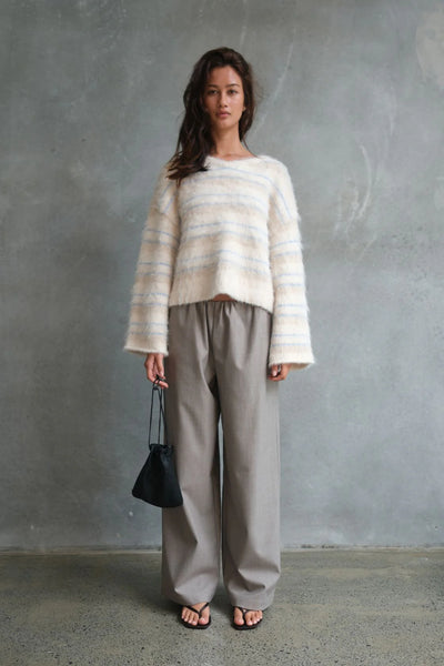 A full-length editorial lifestyle shot featuring a model posing against a textured gray concrete wall. She is wearing the striped Lucca knit sweater paired with taupe wide-leg pants and dark sandals, carrying a minimal black drawstring pouch to emphasize a casual, everyday winter layering look.