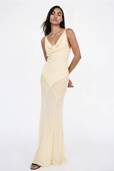 Full-length photo of a model standing with hands behind her back, wearing a butter yellow cowl neck maxi gown with a panelled bias-cut skirt.