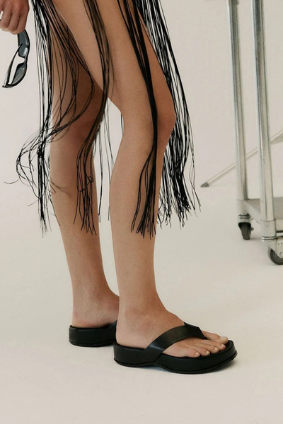 Studio shot focusing on the lower legs of a model wearing the black leather Wallace sandals with a fringe outfit, standing against a neutral background.