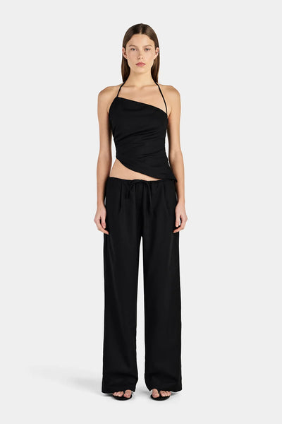 Full-length studio front view of a model wearing a black linen-blend asymmetrical halter top featuring side gathering and an angled hem, paired with relaxed black drawcord pants.