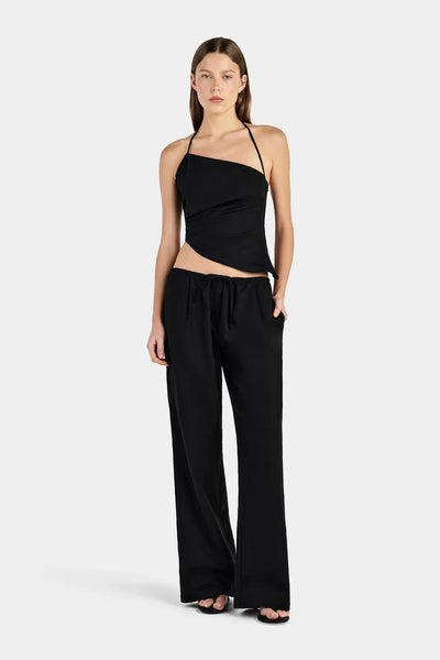 Full-length studio front view of a model with her hand in her pocket, wearing a black asymmetrical halter neck top with side gathering and black straight-leg trousers.